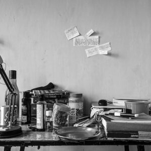 A simple kitchen table with books, bottles, and jars haphazardly piled on it. A desk lamp sits on the corner of the table facing away from the other items.