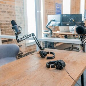 A modern podcast setup with microphones and headphones on a wooden desk in a glass-walled office space featuring exposed brick walls and computer workstations.