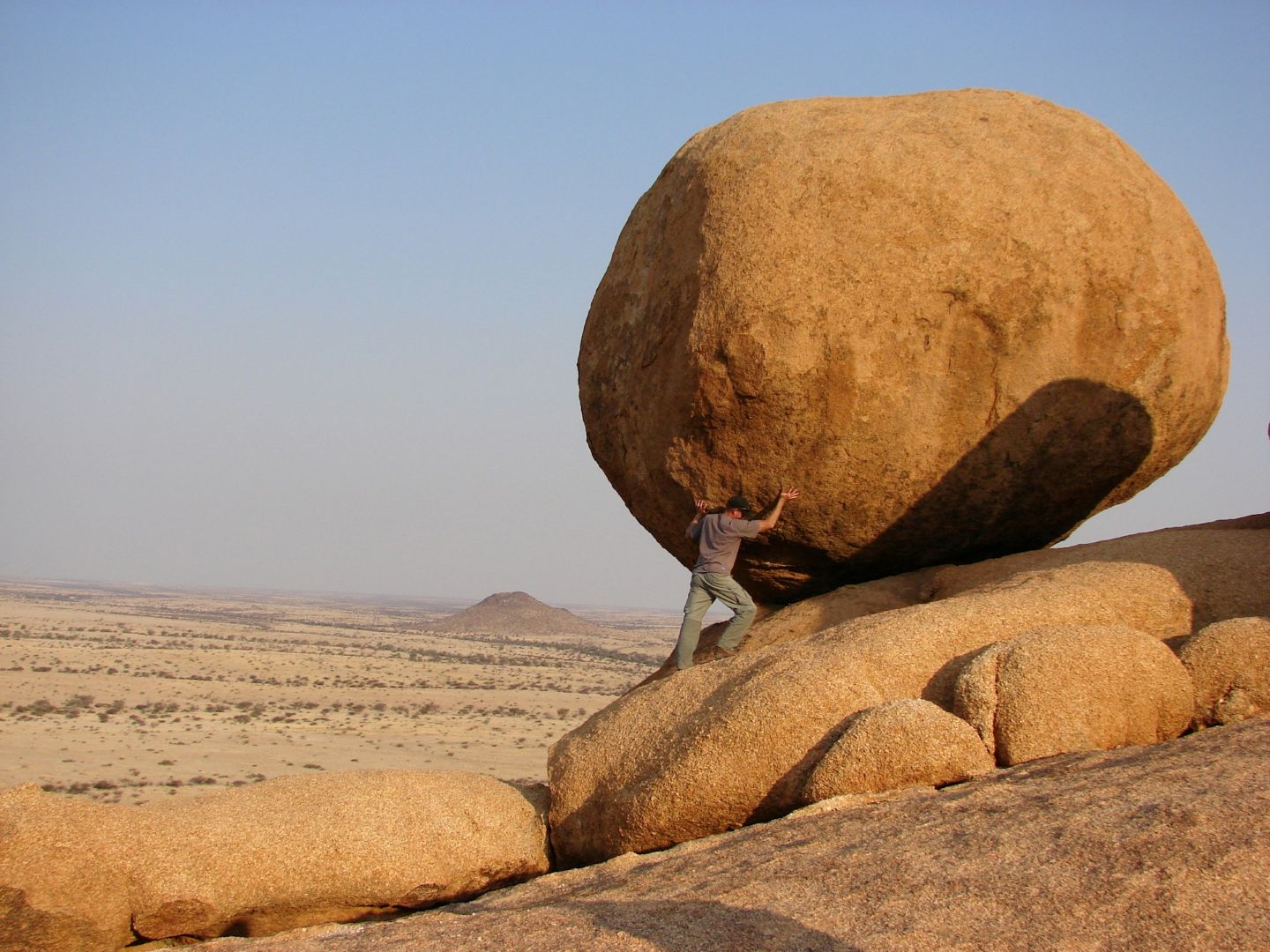 A person pushing against a massive, round boulder balanced on a rocky slope in a dry, open desert landscape. The scene suggests effort, tension, and the scale of the rock compared to the individual.