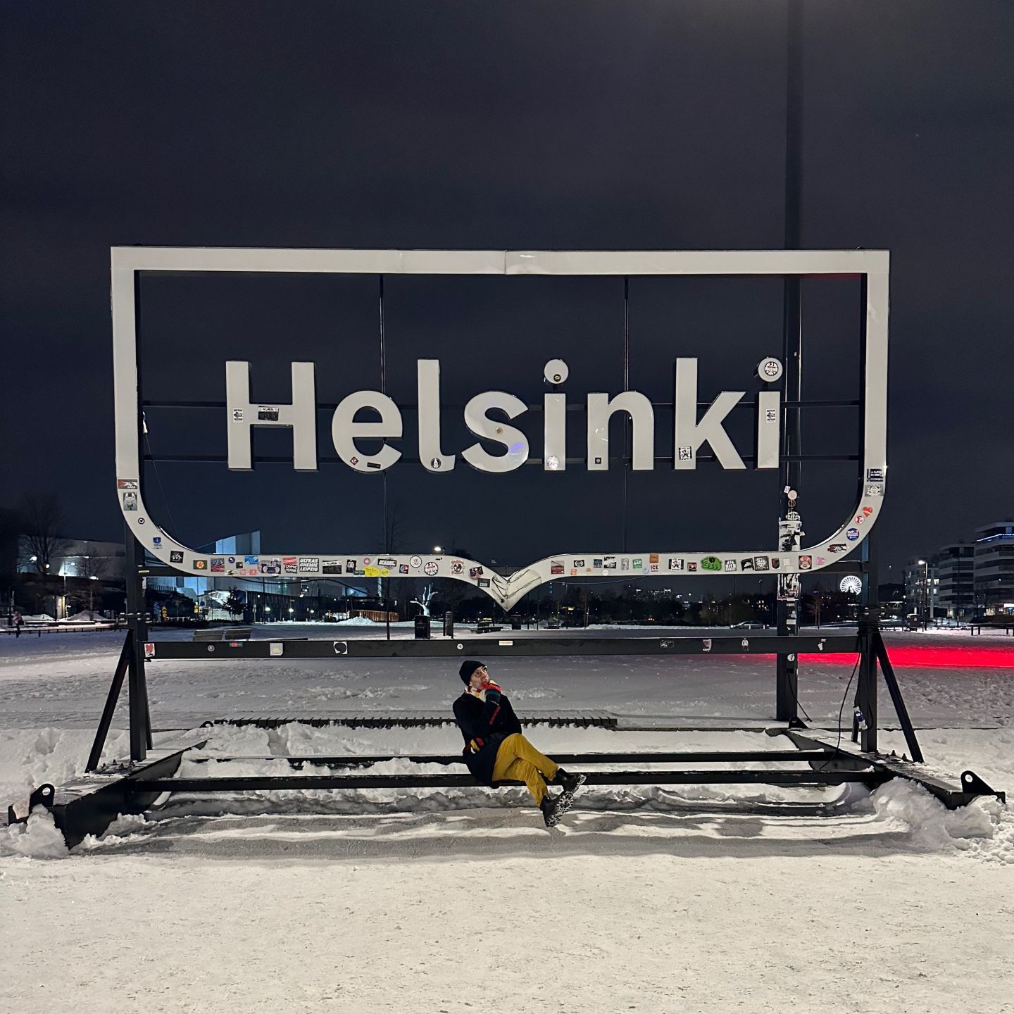 A man sitting under a Helsinki city sign on snow covered ground.