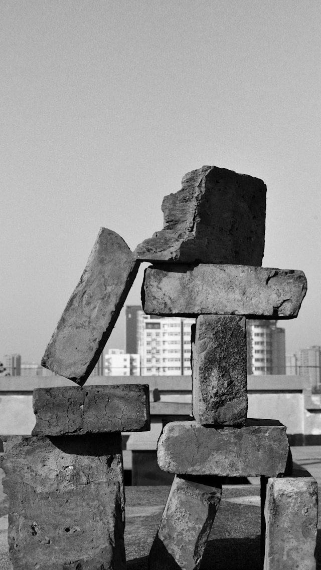 Weathered bricks stacked in a rough, balanced arrangement on a rooftop, with a city skyline visible in the background. Black and white photograph.
