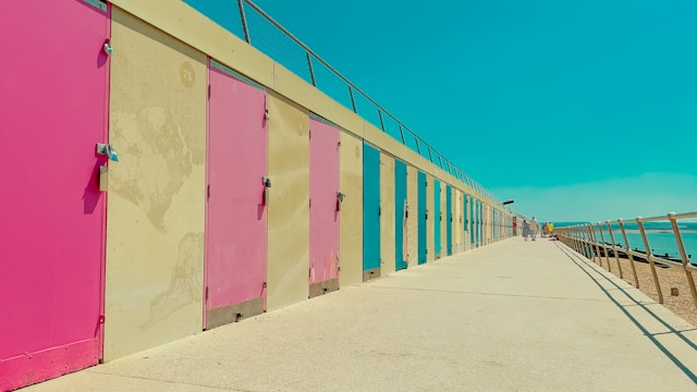 A row of colourful beach huts in pink, cream, and pastel shades lines a seaside promenade under a clear turquoise sky.